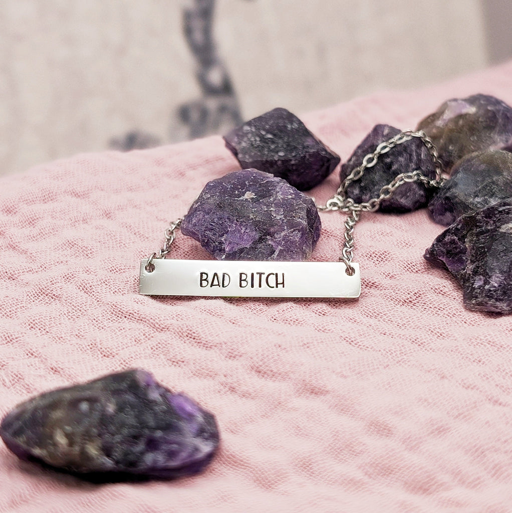 A stainless steel necklace with a bar pendant stamped with the phrase 'BAD BITCH', displayed on a pink fabric with raw purple crystals around it.