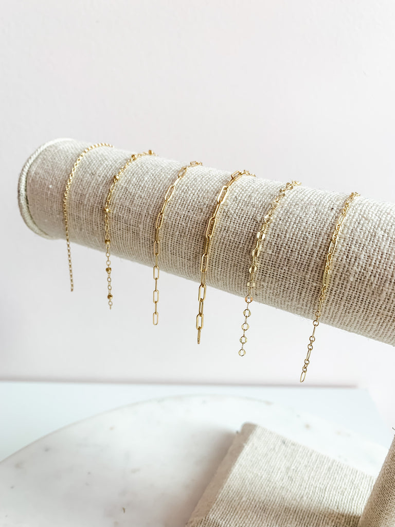 A display of delicate, dainty bracelets made of gold fill, rose gold fill, or sterling silver, laid out on a textured surface.
